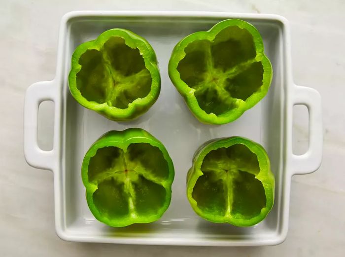 A top-down view of four green bell peppers with their tops removed, resting in a baking dish. 