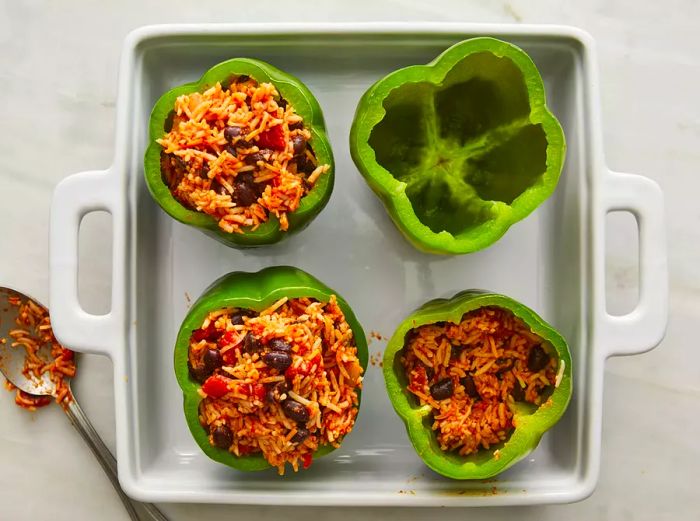 A top-down view of a cheese and rice mixture being spooned into four green bell peppers placed in a baking dish. 