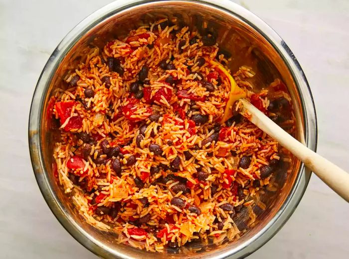 A top-down view of rice, black beans, tomatoes, chili powder, garlic salt, and cumin being stirred together in a pot. 