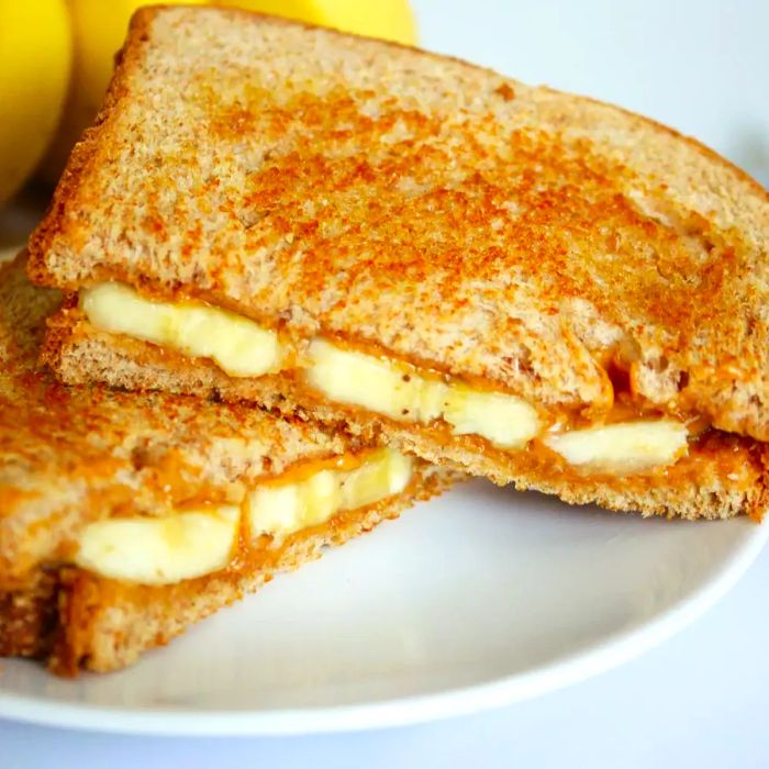 A close-up shot of a Grilled Peanut Butter and Banana Sandwich, sliced in half and placed on a white plate, with a backdrop of bananas.