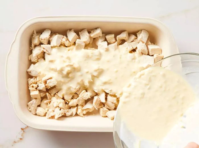 A bowl of creamy soup and milk mixture being poured over chicken pieces in a baking dish