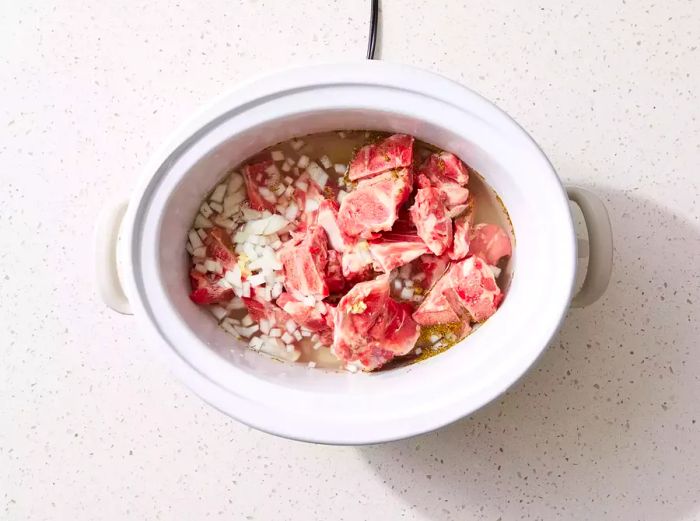 A slow cooker filled with neck bones, onion, garlic, thyme, salt, and vinegar.