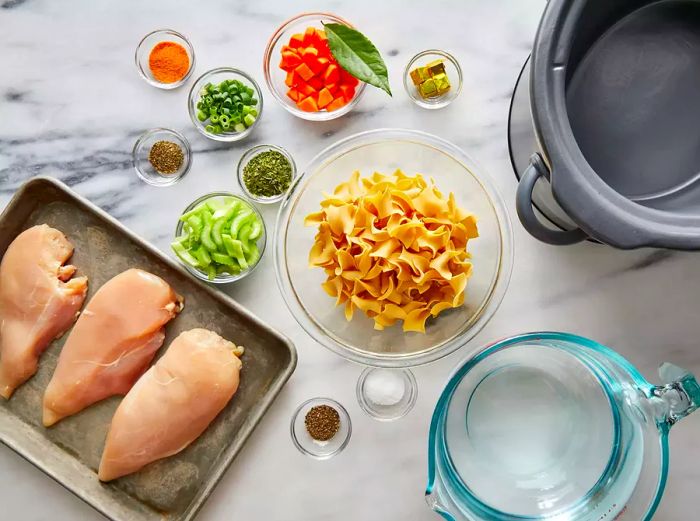 Aerial view of chicken noodle soup ingredients arranged in different bowls and pans.