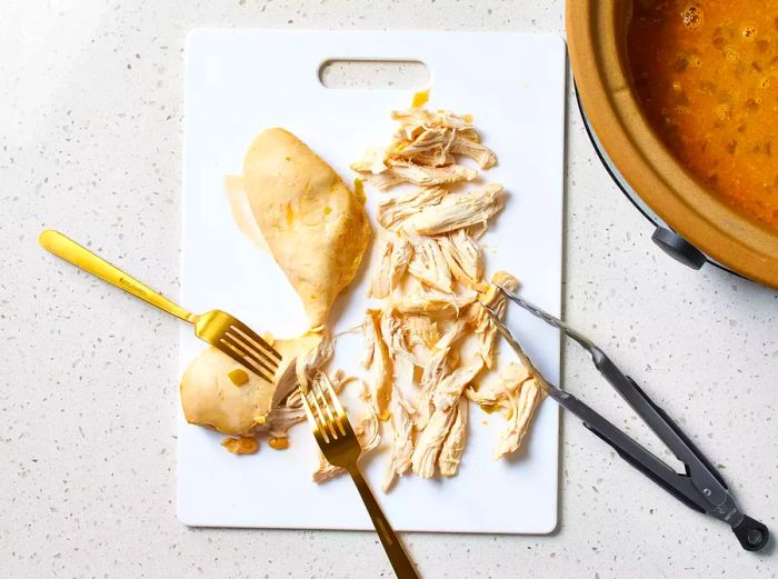 A cutting board with chicken breasts being shredded with forks.