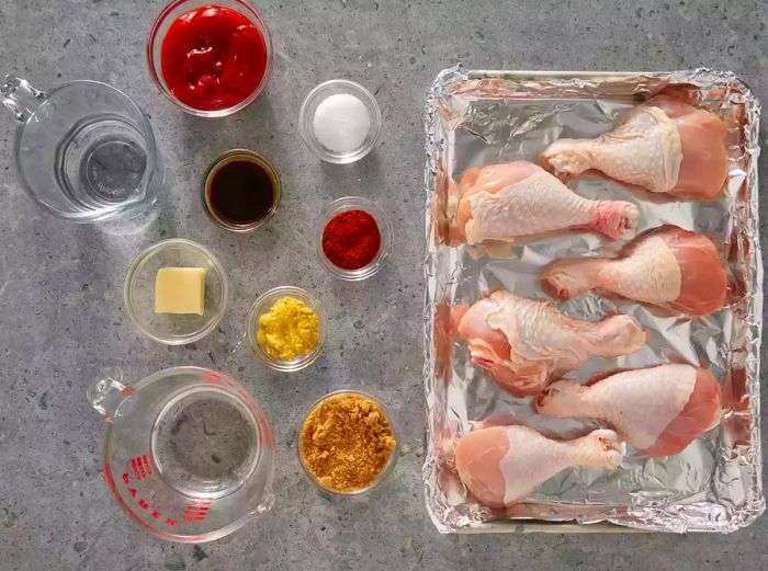 Aerial view of ketchup, vinegar, water, brown sugar, butter, salt, Worcestershire sauce, mustard, and chili powder in glass bowls, along with five raw drumsticks.