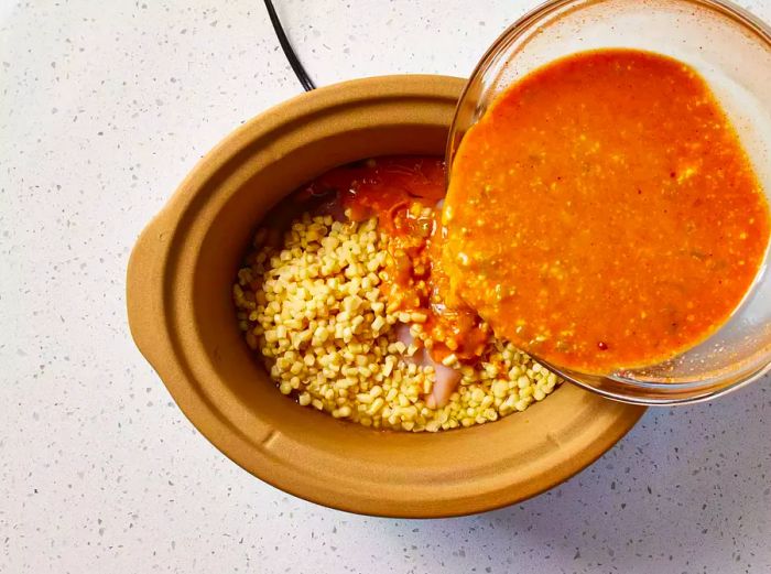 A slow cooker with a mixture of condensed soup and chiles being poured over chicken breasts, beans, and corn.