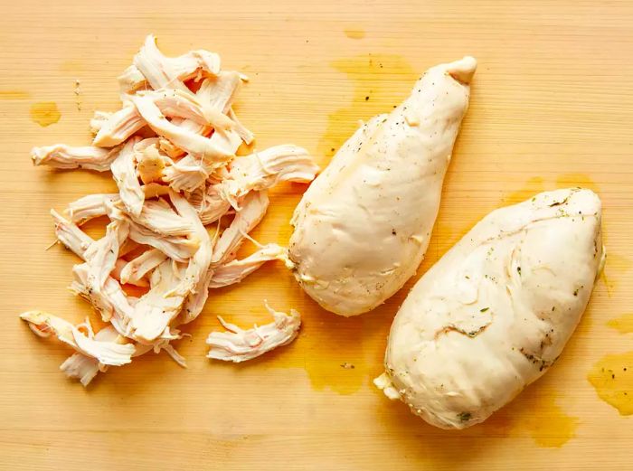 Overhead shot of shredded chicken alongside two whole chicken breasts resting on a wooden surface.