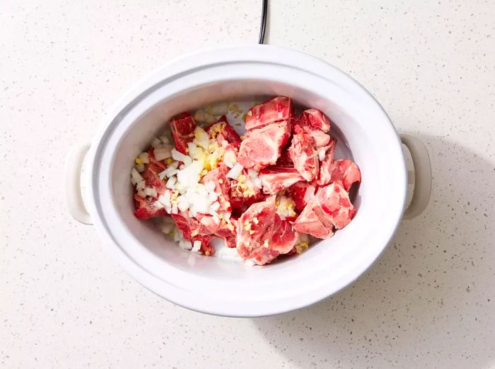 A slow cooker filled with neck bones, onion, and garlic.