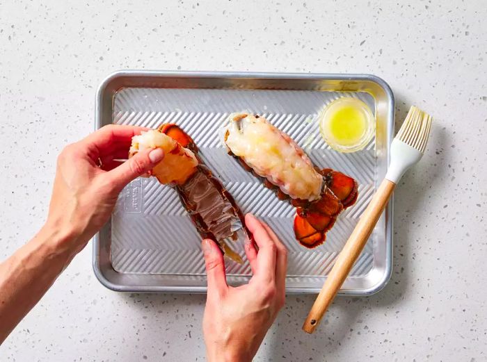 Hands gently extracting a lobster tail from its shell on a baking sheet