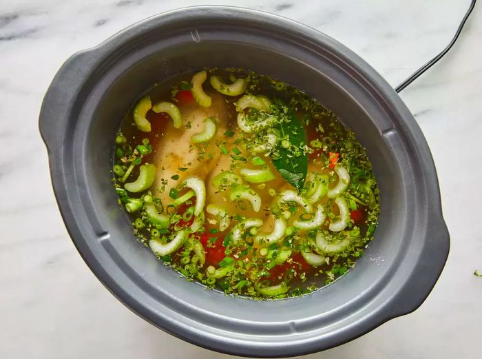 Aerial view of chicken breasts, broth, chopped vegetables, and spices placed in a slow cooker.