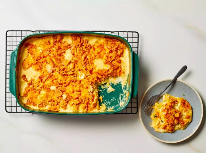 A plate of Million Dollar Potato Casserole, topped with fresh thyme, served alongside a fork and a baking dish resting on a cooling rack.