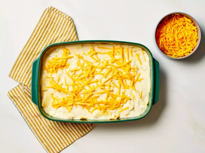 Potatoes and creamy chicken soup mixture layered in a baking dish, topped with cheese, next to a bowl of extra cheese.