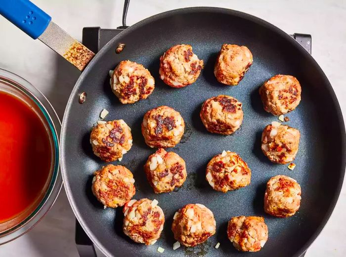 Meatballs cooking in a skillet, with a bowl of sauce placed to the side.