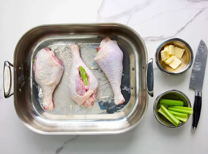 A bird's-eye view of three raw turkey legs, coated in butter with celery stuffed inside, resting in a baking dish.