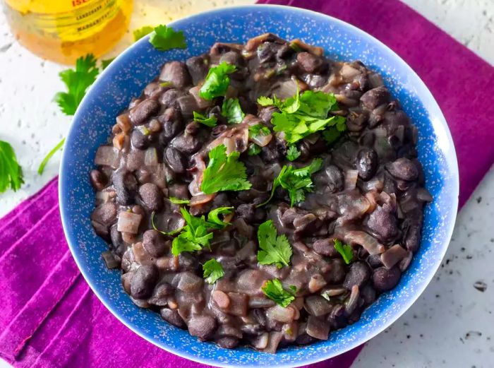 An overhead shot of a bowl of black beans topped with herbs, placed on a blue bowl and a purple kitchen towel.