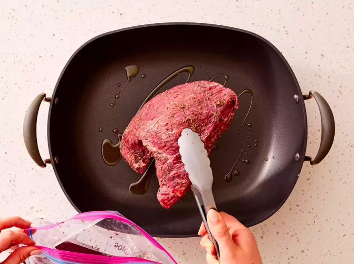 Using tongs, add the marinated tri-tip beef to the oiled roasting pan.