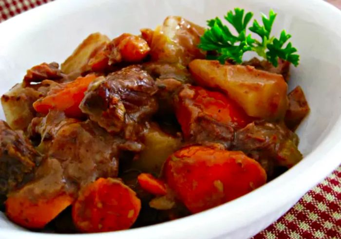 A close-up of a white ceramic bowl filled with beef stew made from carrots, potatoes, and onions, topped with a sprig of parsley