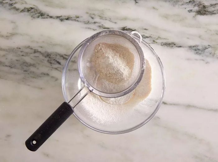 Aerial view showing flour, garlic salt, onion salt, paprika, and salt being sifted into a mixing bowl.