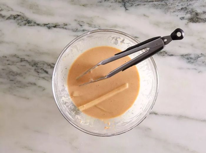 French fries being dipped into a batter inside a glass bowl.