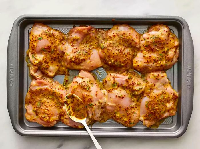 A top-down shot of chicken thighs resting on a baking sheet, with mustard sauce being spooned on top.