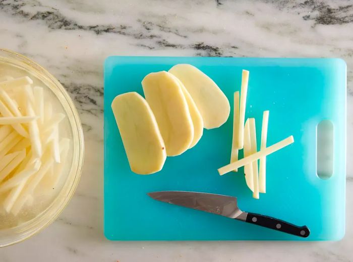 Aerial view showing potatoes being sliced into fries and submerged in a bowl of cold water.