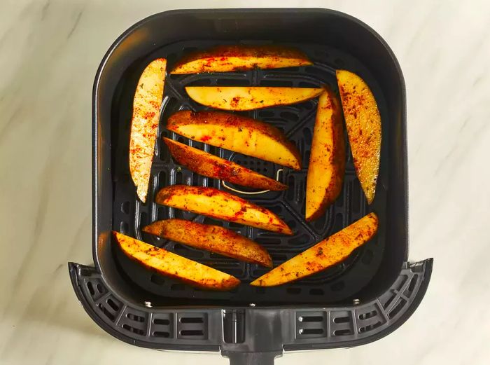 An overhead shot of the seasoned potato wedges arranged in the basket of an air fryer.