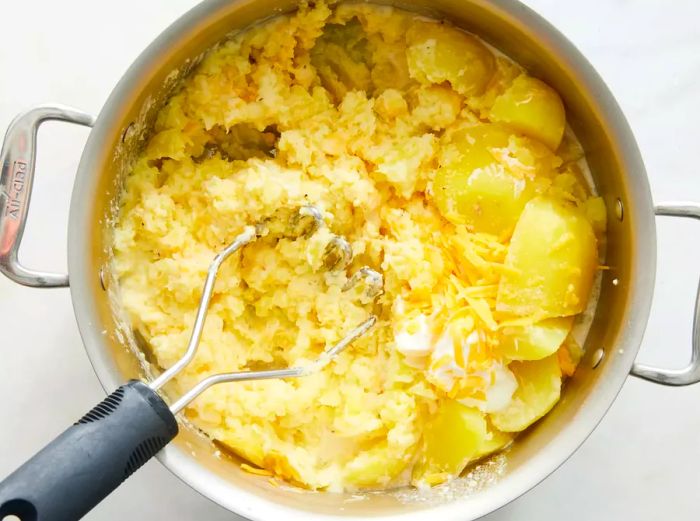 Creamy mashed potatoes mixed with cheese and sour cream in a pot, being mashed with a potato masher