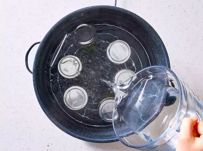 A hand pouring hot water from a pitcher into a large stockpot of simmering water with a rack, holding six jars of green beans