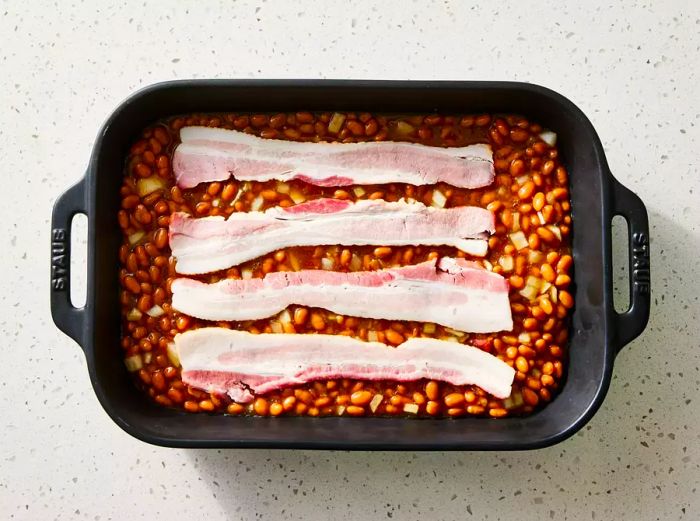 A baking dish filled with seasoned beans, topped with four slices of raw bacon.