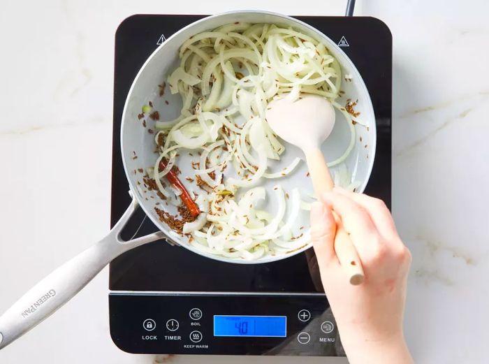 A pot with toasted spices and sliced onion being stirred with a spatula