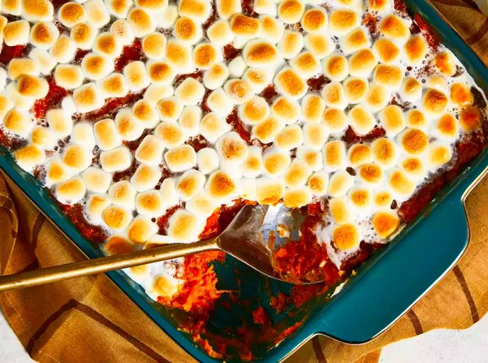 A casserole dish filled with sweet potatoes topped with golden, toasted marshmallows, with a hearty scoop taken out