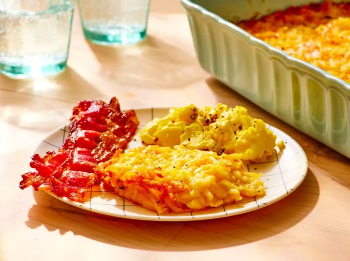 A plate of crispy restaurant-style hashbrown casserole paired with scrambled eggs, and a baking dish of the casserole in the background.