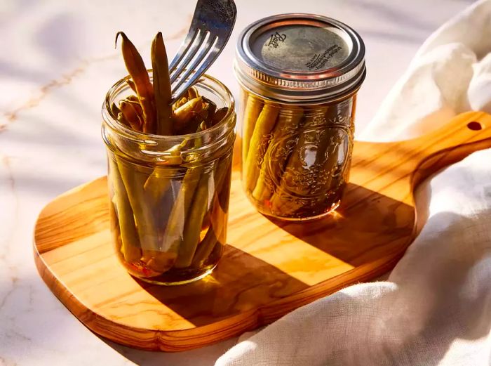 Two jars of crisp pickled green beans resting on a cutting board, with a fork lifting green beans from an open jar