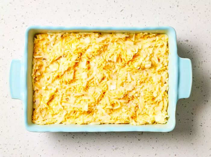 A greased casserole dish filled with shredded potatoes, creamy soup, cheese, onion, butter, and seasoned with salt and pepper.