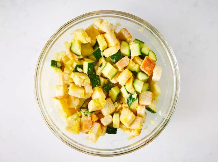 A bird's eye view of cubed bread, zucchini, onion, egg, and garlic salt in a glass mixing bowl