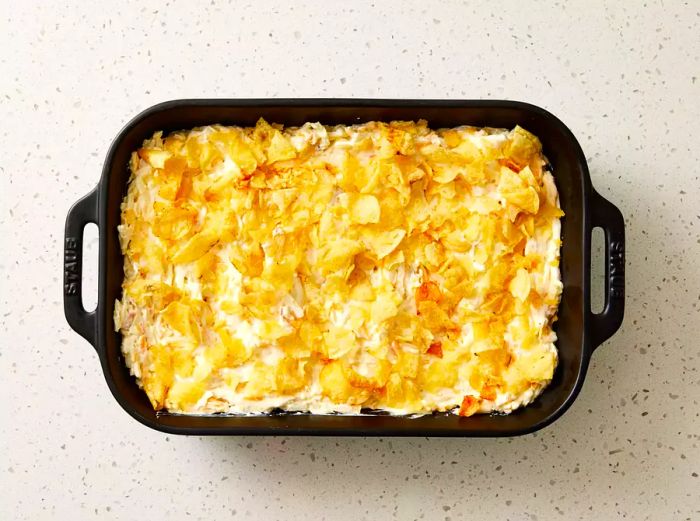 A casserole dish filled with the hash brown and sour cream mixture, topped with crushed potato chips.