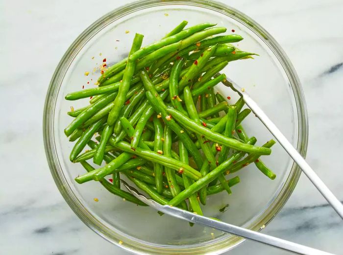 An overhead view of the marinated green beans in a bowl, being tossed with tongs