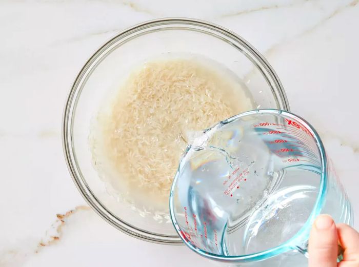 A hand pouring a measuring cup of water into a glass bowl filled with basmati rice