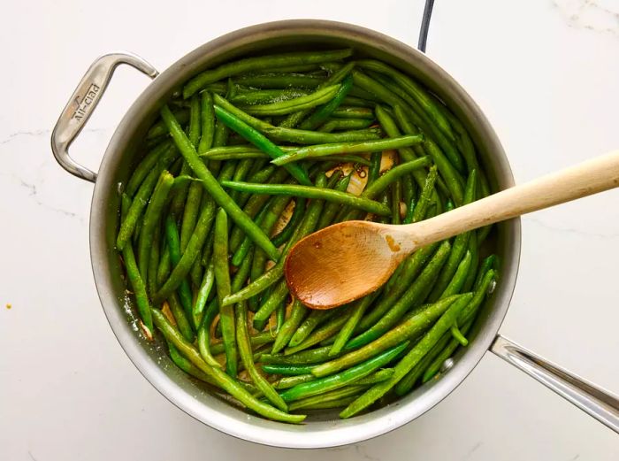 A wooden spoon stirring green beans in a pan with soy sauce, oyster sauce, and sugar