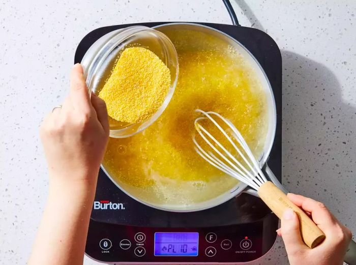 A hand pouring a small bowl of polenta into boiling water, while another hand stirs the mixture to ensure smoothness.