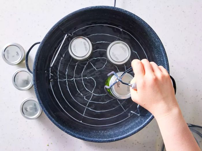 A jar holder gently lowering a sealed jar of green beans into simmering water in a stockpot with a rack