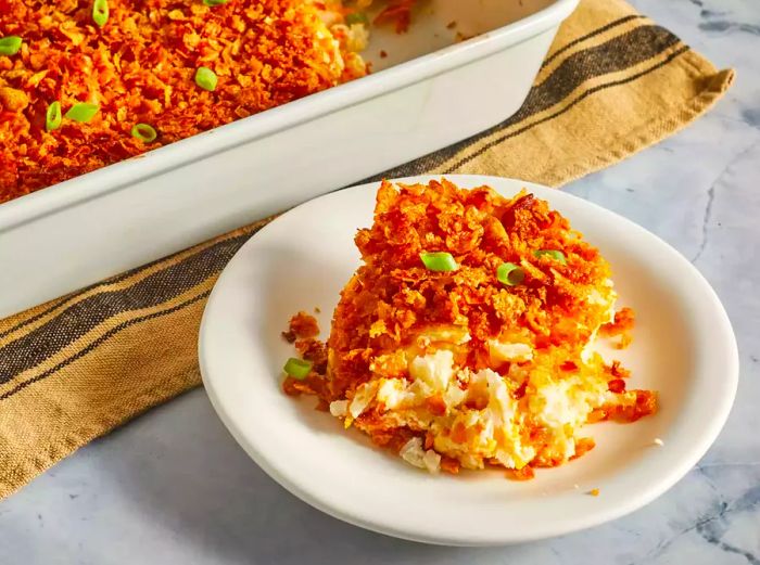 A low-angle, close-up shot of a generous scoop of cheesy potato casserole plated in front of a casserole dish, with the remaining casserole garnished with fresh green onions.