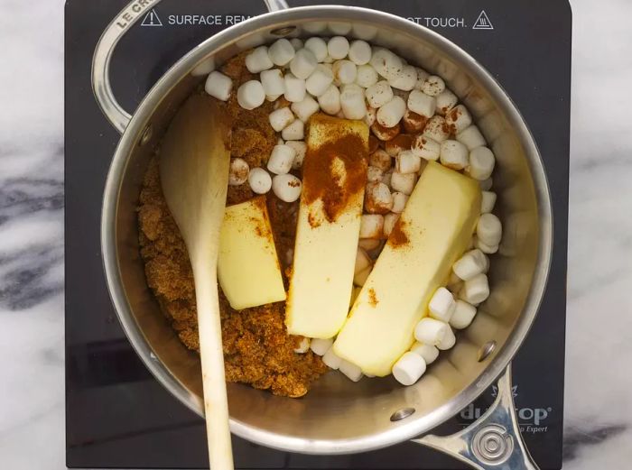 A large pot containing two sticks of butter, marshmallows, brown sugar, cinnamon, and nutmeg, being stirred with a wooden spoon