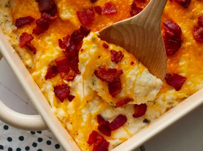 A high-angle close-up of a baking dish filled with twice-baked potato casserole topped with bacon, being spooned out.