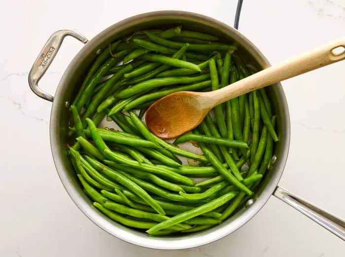 A wooden spoon stirring green beans in a large skillet