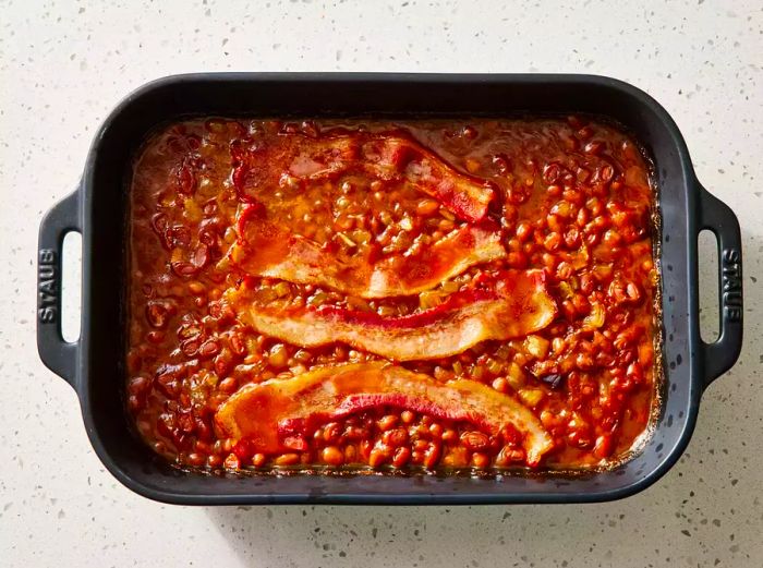 A baking dish of baked beans with crispy bacon slices on top.