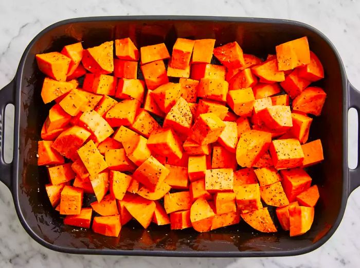 An overhead shot of diced sweet potatoes in a baking dish, seasoned with oregano, salt, and pepper.