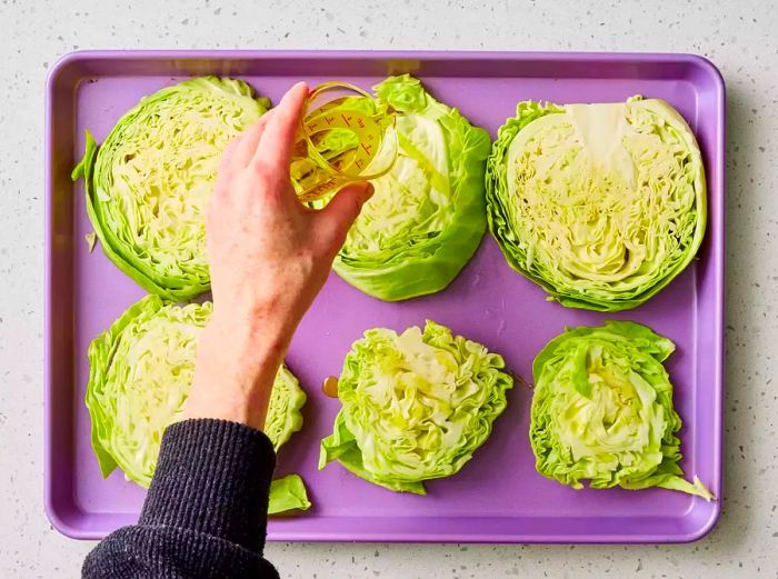 A baking sheet with six thick cabbage slices being drizzled with olive oil