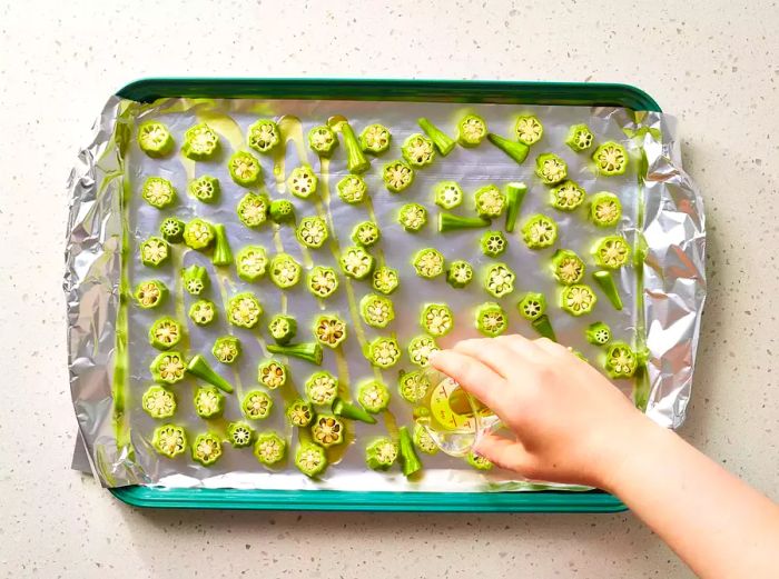 A baking sheet lined with aluminum foil, topped with sliced okra and drizzled with olive oil