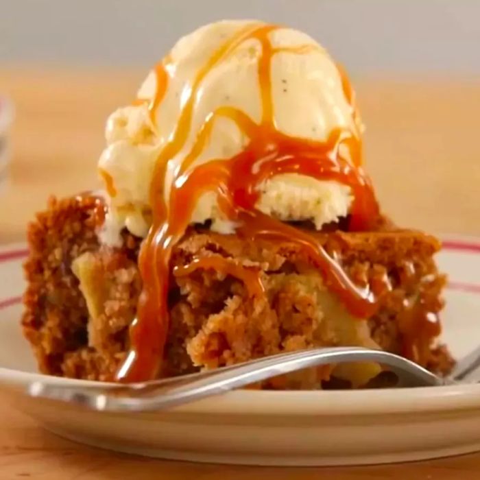 A close-up of a slice of Fresh Apple Walnut Cake, topped with a scoop of ice cream and drizzled with caramel, served with a fork on the side.